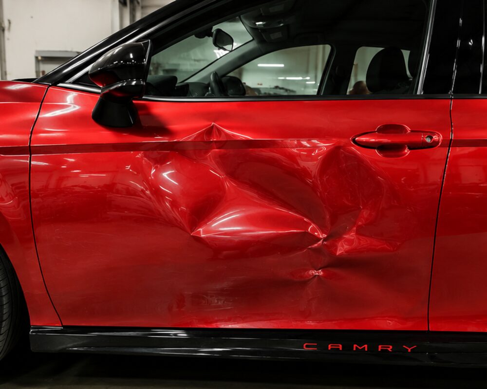 Red Toyota Camry with a deep impact dent through the driver door and rocker panel, before repair